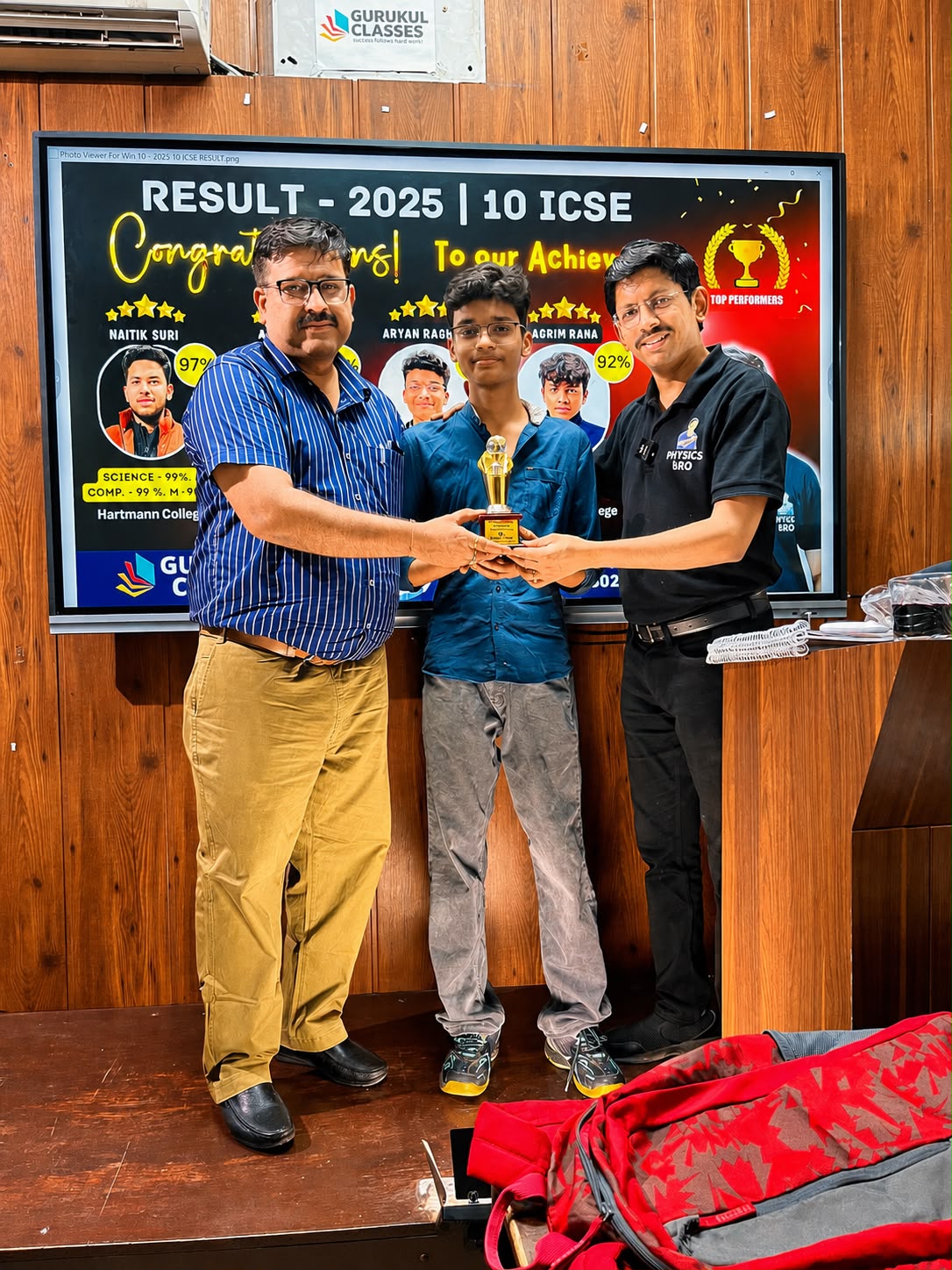Class 10 ICSE 2025 achiever receiving a trophy in front of the results board at Gurukul Classes