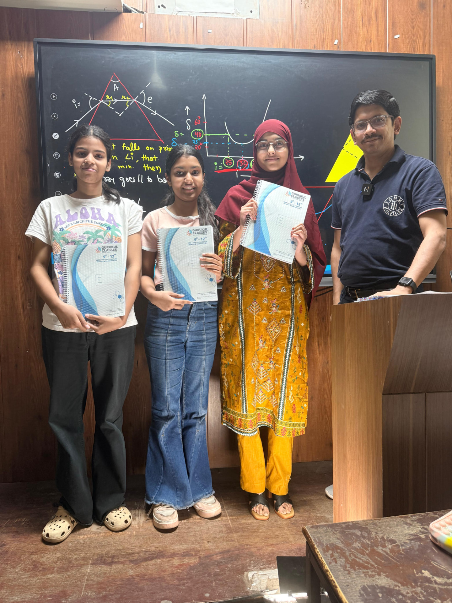 Students holding Gurukul Classes notebooks in front of an optics smart-board lesson