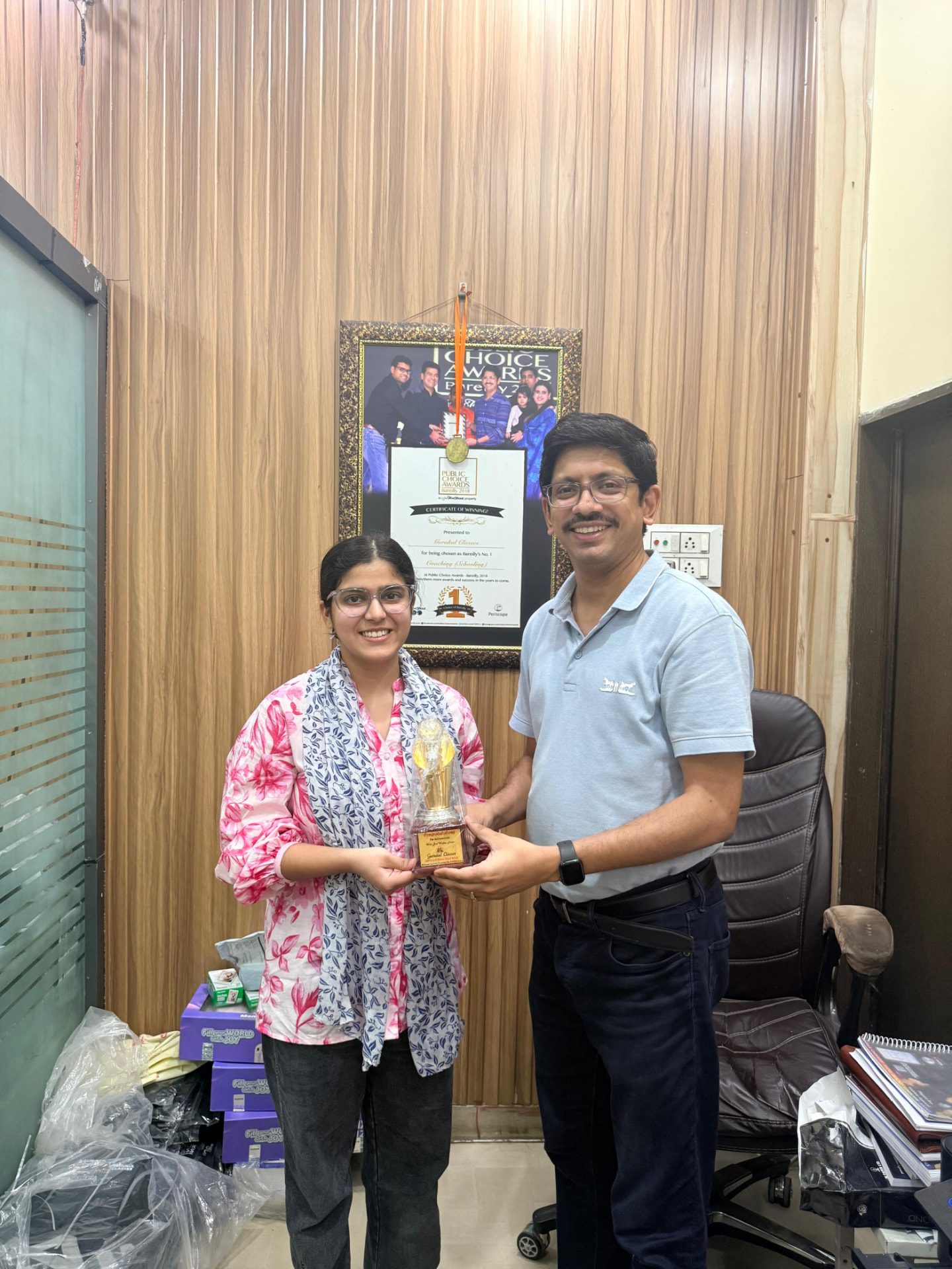Achiever receiving a Gurukul Classes trophy at the institute office beside the Public Choice Awards certificate